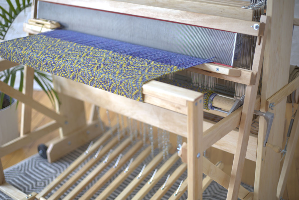 A floor loom showing an in-progress blue and yellow patterned weaving project